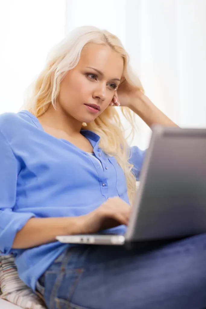 A woman with long blonde hair sits relaxed, wearing a blue shirt and jeans, looking thoughtfully at a laptop screen as she types. The background is softly lit.