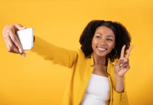 A smiling woman wearing a yellow blazer takes a selfie with her smartphone, holding up a peace sign with her other hand, against a solid yellow background.