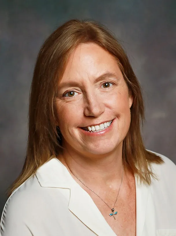 A woman with straight, light brown hair wears a white blouse and a dragonfly necklace, smiling at the camera against a soft, dark background.