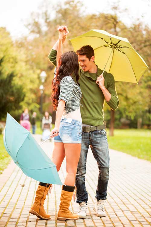 A smiling couple holding umbrellas dances together on a park pathway. The woman wears a checkered shirt, shorts, and boots, while the man wears a green sweater and jeans. Trees and greenery are visible in the background.