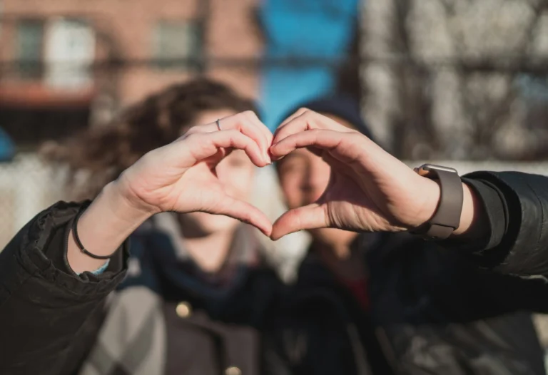 Two people stand close together outdoors, forming a heart shape with their hands in the foreground. Their faces are blurred, and a building and blue sky are visible in the background.