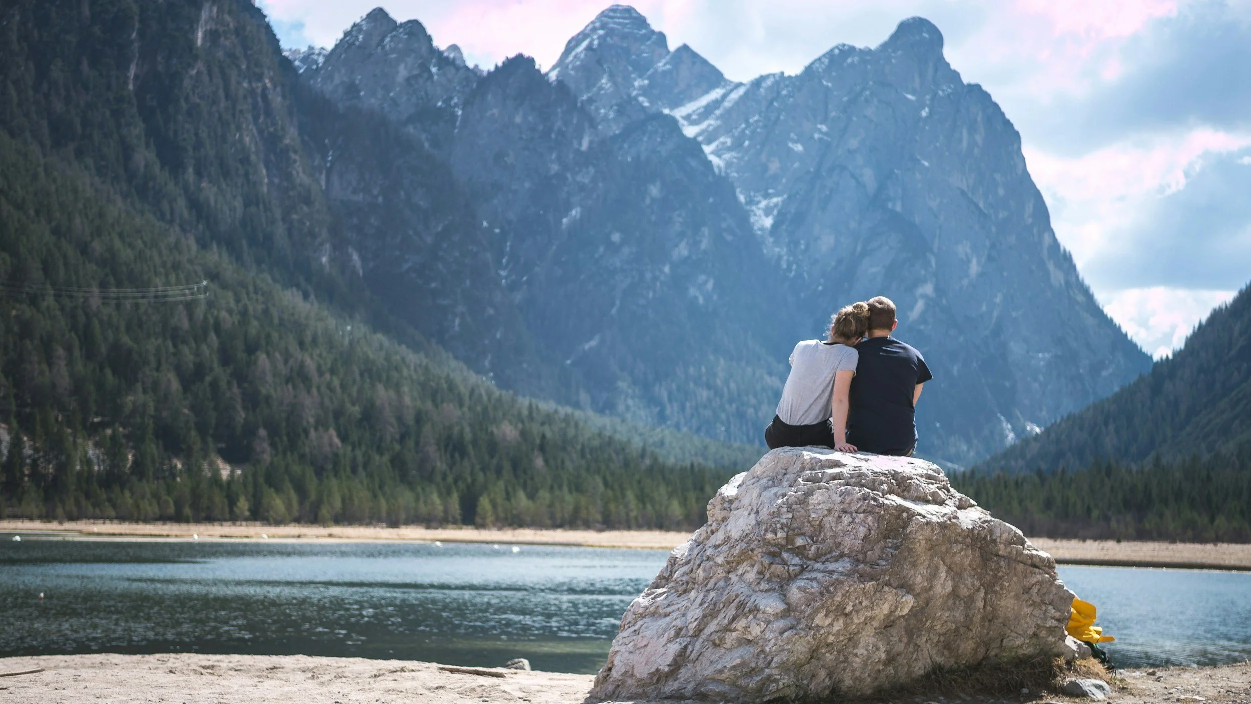 A couple sits closely together on a large rock by a calm lake, surrounded by dense pine forests and tall, rugged mountains under a partly cloudy sky.