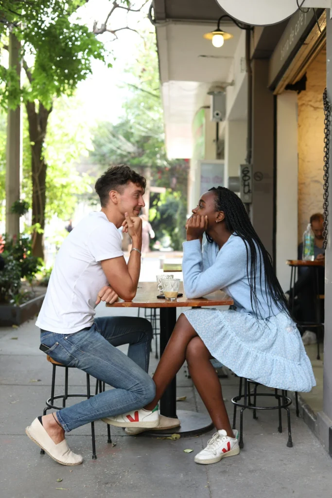 A young couple sits at a small outdoor café table, smiling and leaning toward each other while talking. The woman wears a blue dress and the man wears a white shirt and jeans. Trees and shops line the street in the background.