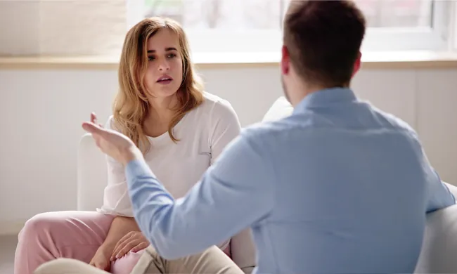 Two people sit indoors having a serious conversation. The woman looks concerned while listening, and the man gestures with his hand as he speaks. Natural light comes through a window behind them.