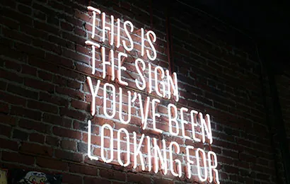 A neon sign on a brick wall reads, "THIS IS THE SIGN YOU'VE BEEN LOOKING FOR" in bright white letters.