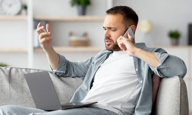A man sitting on a couch with a laptop on his lap, talking on his smartphone and looking frustrated, gesturing with his hand as if confused or upset.