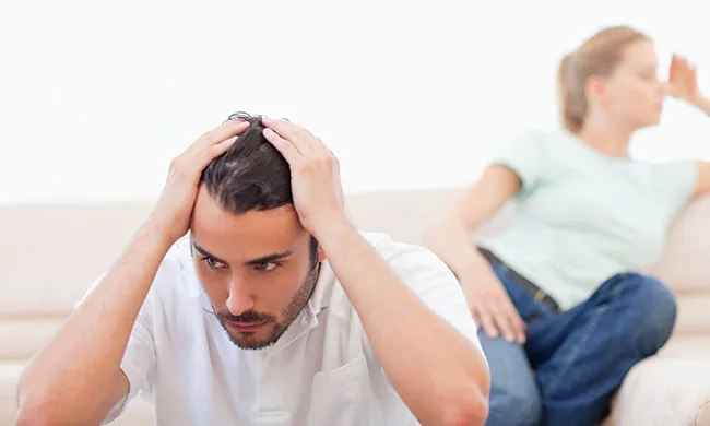 A man sits on a couch holding his head in distress, while a woman sits behind him, turned away with an upset expression. Both appear to be troubled, suggesting a tense or emotional situation.